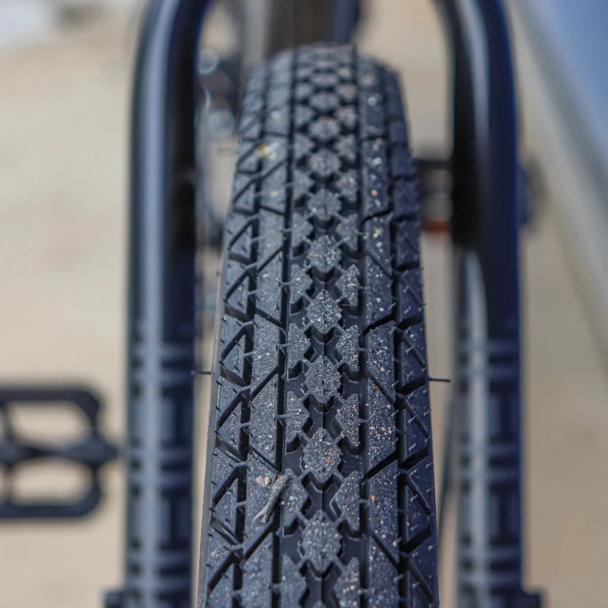 Black knobby tread bicycle tire close-up – Close-up of a black commuter bicycle tire showing a deeply grooved diamond-pattern tread with scattered light grit, framed between the blurred rear fender and seat stay of the bike.