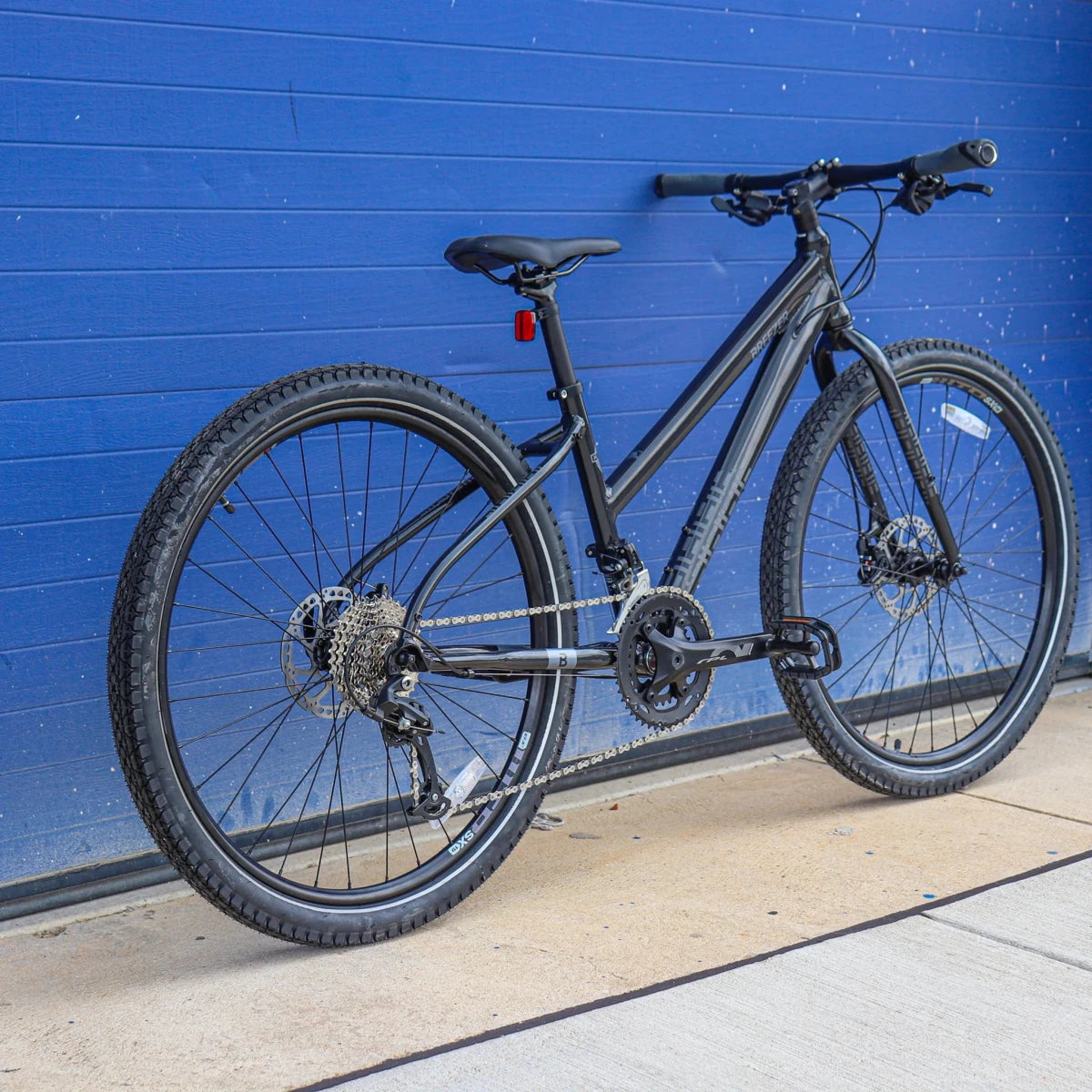 Black Breezer Midtown 1.5 ST step-through hybrid bicycle – Matte black step-through hybrid bike with flat handlebars, knobby tires, hydraulic disc brakes, and a rear cassette derailleur leaning against a bright blue paneled wall on concrete pavement.