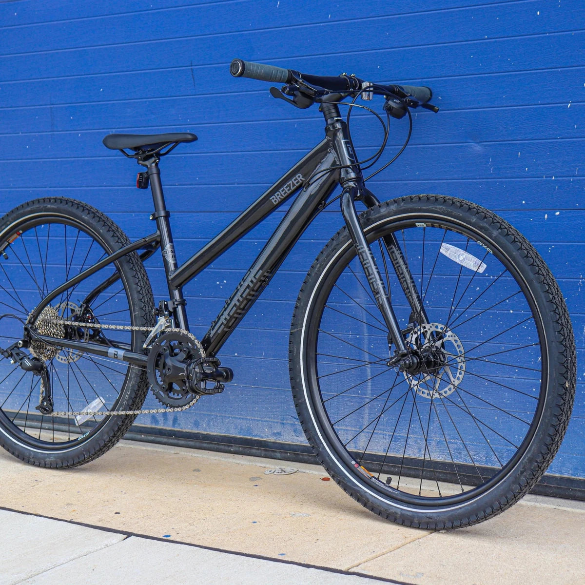 Black Breezer Midtown 1.5 ST hybrid bicycle – A matte black step-through hybrid bicycle with flat handlebars, knobby tires, and disc brakes leans against a bright blue corrugated metal wall on a concrete surface, displaying the 'Breezer' brand on the down tube.