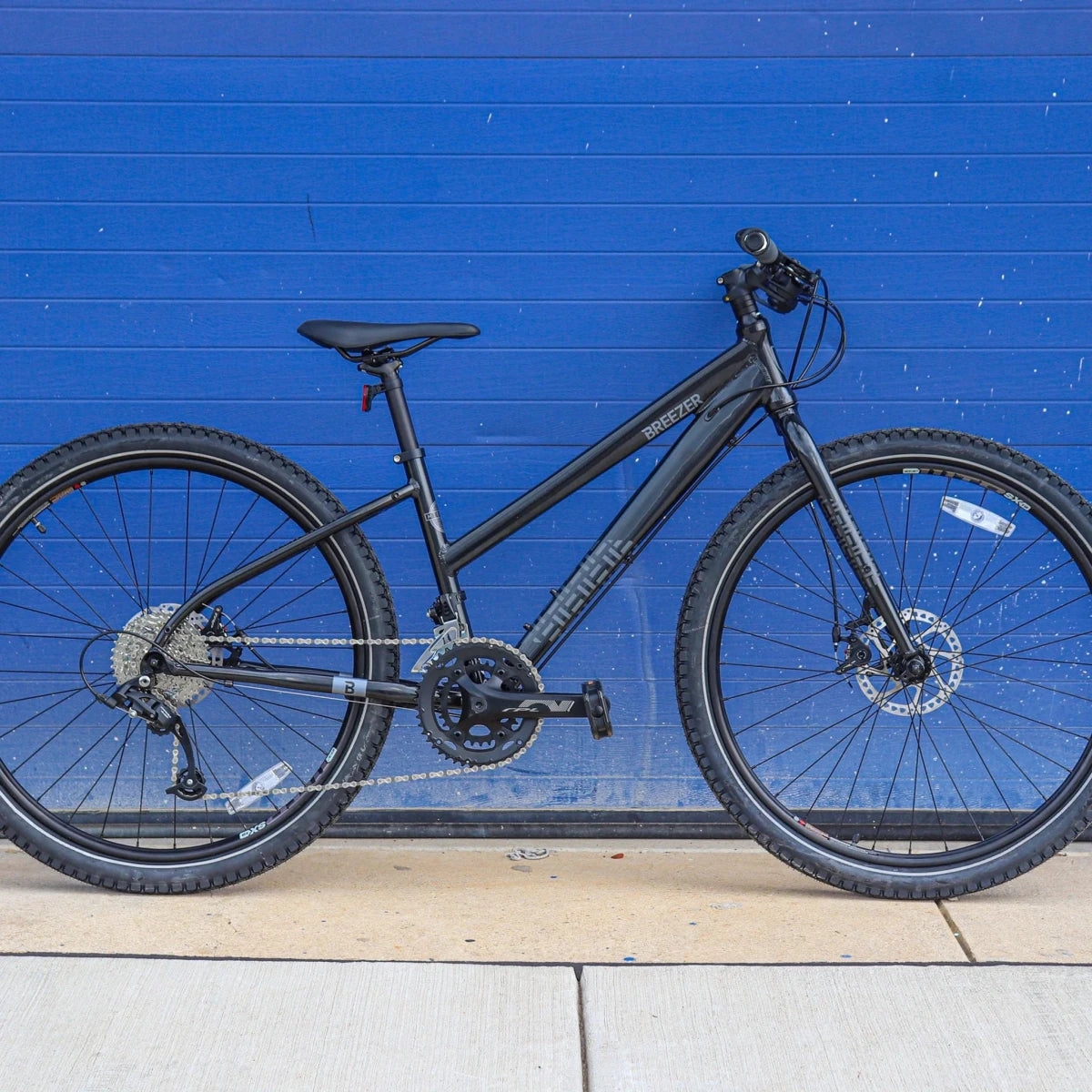 Black Breezer Midtown step-through hybrid bicycle – Dark grey step-through Breezer hybrid bike with flat handlebars, Shimano drivetrain, and hydraulic disc brakes, posed in profile against a vivid blue paneled wall on a concrete sidewalk.