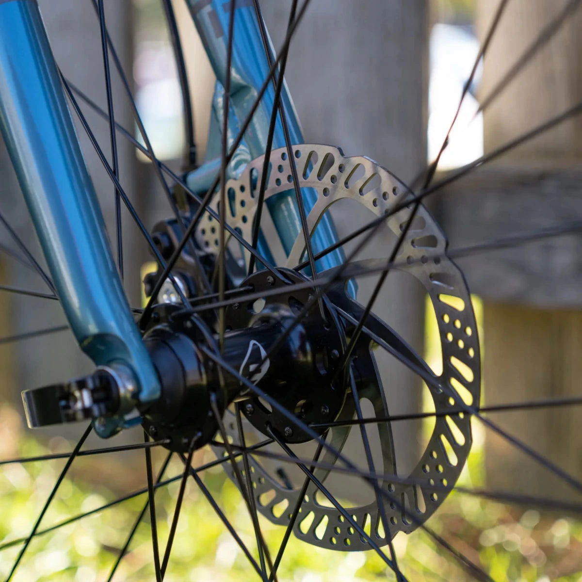 Breezer Midtown hydraulic disc brake rotor – A close-up shot of a bicycle's front wheel hub, showcasing a silver steel disc brake rotor mounted on a black hub with thin spokes, set against a blue fork.