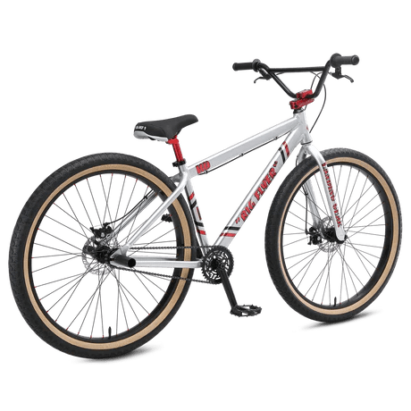 SE Big Flyer HD cruiser bicycle in silver – Silver aluminum wheelie cruiser bicycle shown at a three-quarter angle on a white background, featuring red graphics, black disc brakes, and tan sidewall tires.