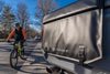 Black Kuat Huk half tailgate pad – A durable black tailgate pad covers half a truck tailgate with secure straps, as a cyclist rides a mountain bike nearby on a sunny day.
