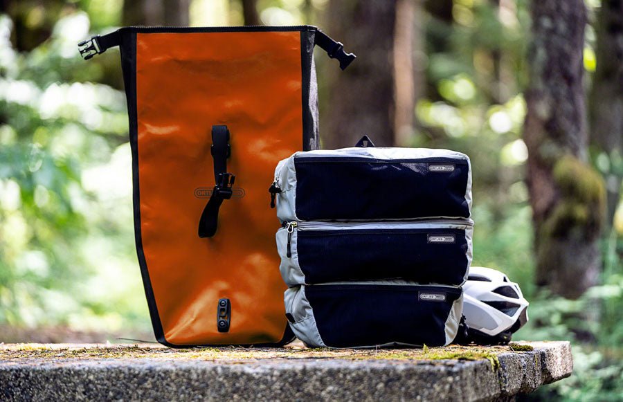 Gray Ortlieb packing cubes stacked outdoors – Three zippered gray packing cubes with black mesh panels stacked next to an orange Ortlieb pannier bag and bike helmet on an outdoor stone table.