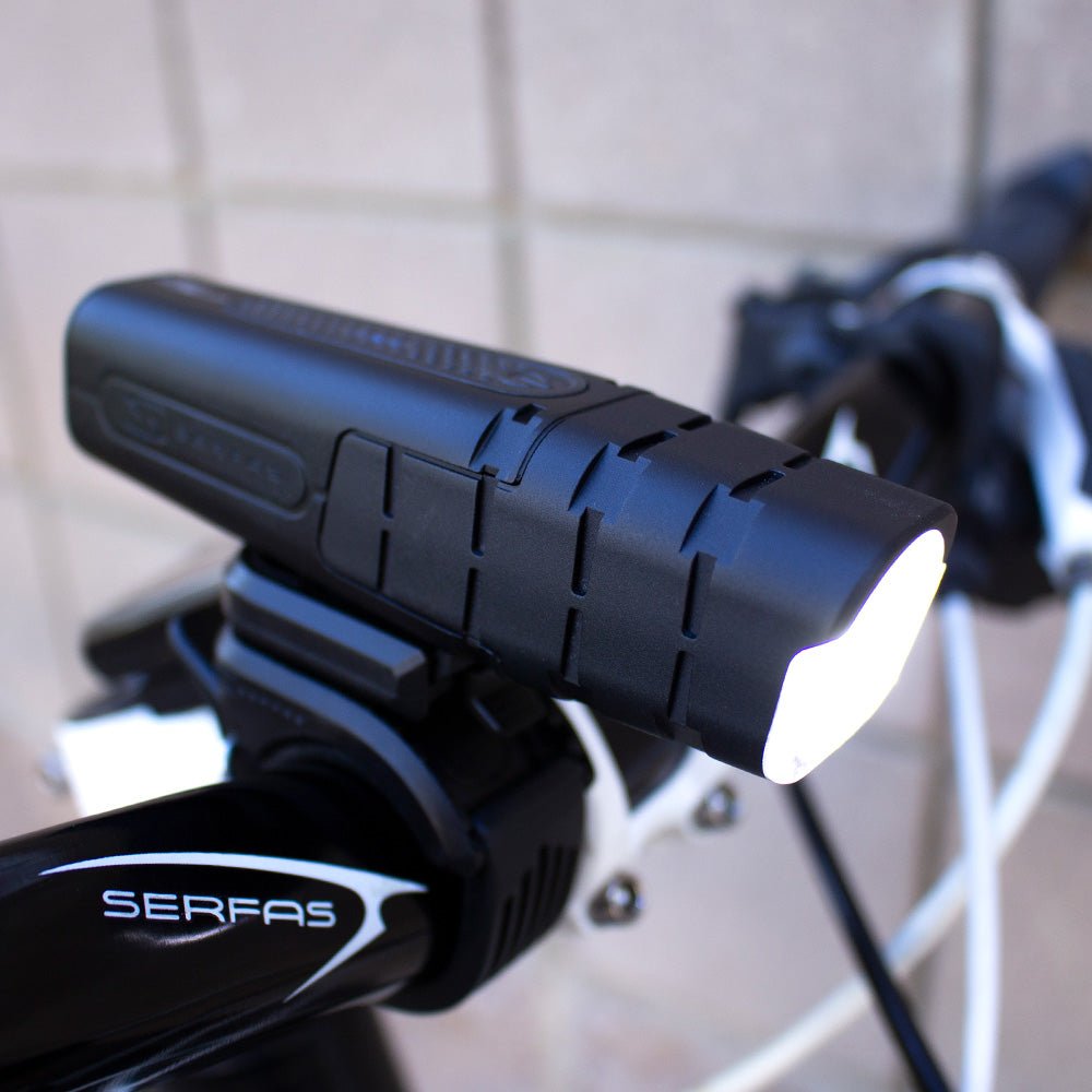 Serfas True 600 MTB handlebar headlight – A matte black Serfas True 600 bicycle headlight with textured grooved casing glows bright white while mounted onto black handlebars against a light wall.