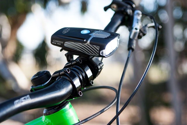 NiteRider Lumina Dual 1800 bicycle headlight – Mounted to the black handlebar of a green bicycle, this compact black and silver LED headlight shines brightly outdoors against a blurred natural background.