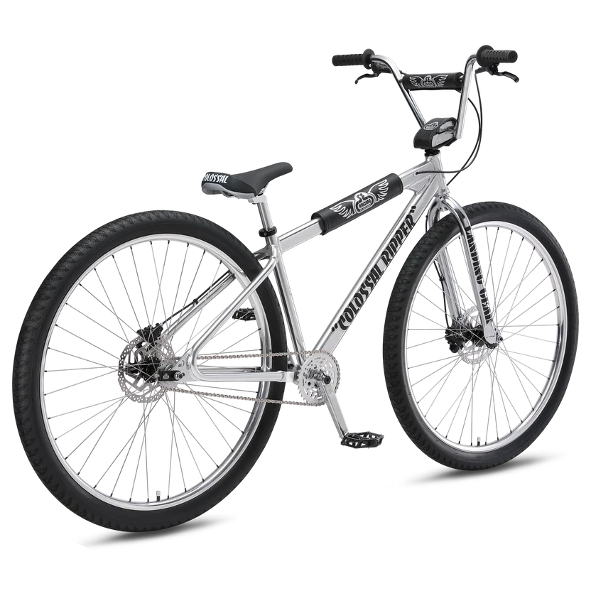 SE Colossal Ripper 32 silver BMX bike – A polished chrome-silver BMX cruiser bike stands on a white background, featuring large 32-inch black knobby tires, dual disc brakes, and retro black frame pads.