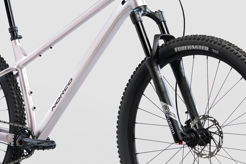 Norco Charger 2 mountain bike front frame – Close-up of a silver Norco hardtail mountain bike featuring an X-Fusion suspension fork, disc brake, and knobby Forekaster front tire against a light gray background.