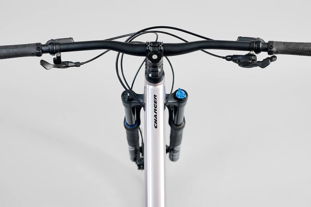 Norco Charger mountain bike handlebar cockpit – Top-down view shows black riser handlebars with rubber grips, brake levers, and shifters mounted to a silver frame with black suspension forks against a light grey background.