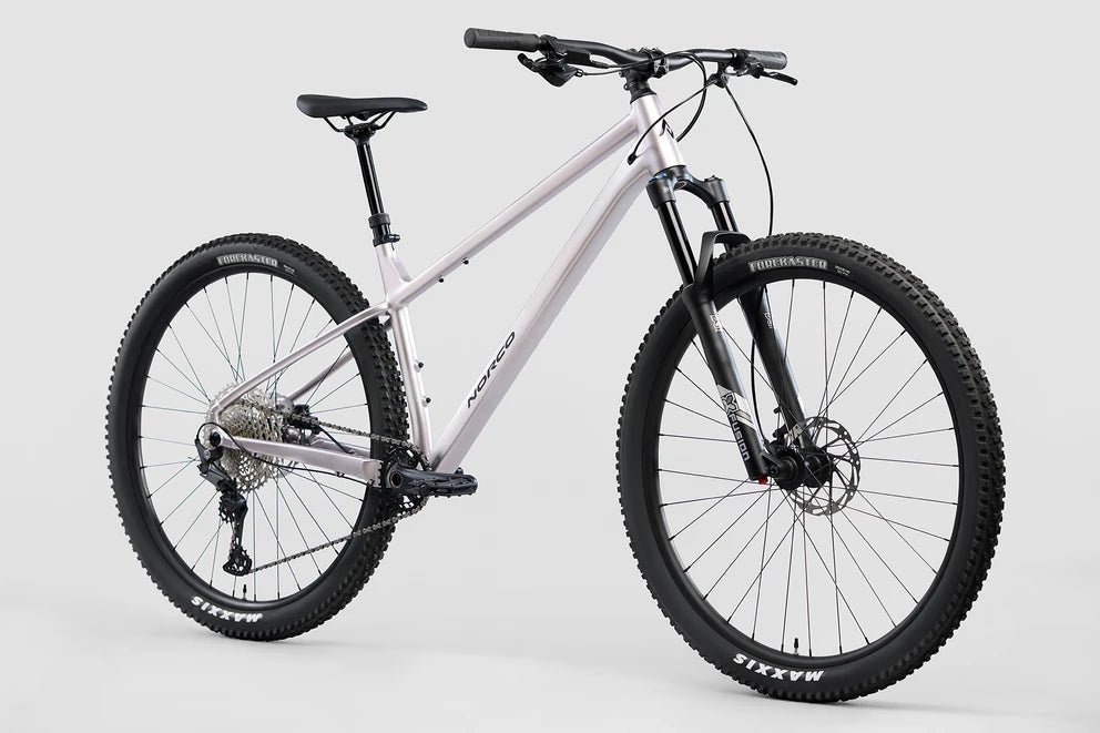 Norco Charger 2 hardtail mountain bike – This silver-lavender hardtail mountain bike features a front suspension fork, knobby black Maxxis tires, disc brakes, and a black saddle, shown at a three-quarter angle against a light gray background.