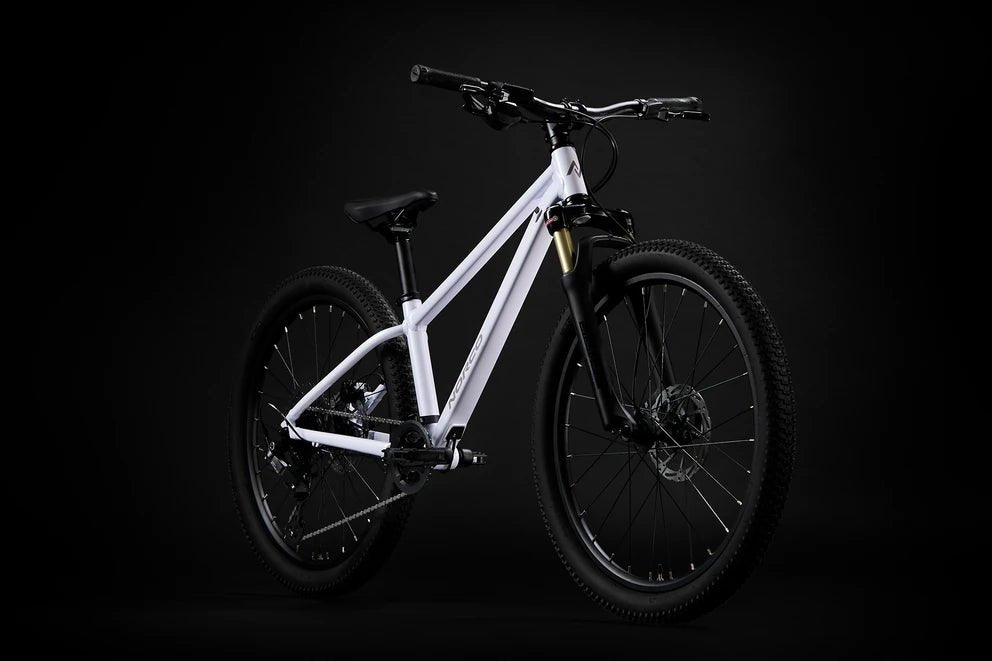 White Norco Charger 24 youth mountain bike – A white youth mountain bike with black knobby tires, suspension fork, and disc brakes stands angled against a solid dark studio background.