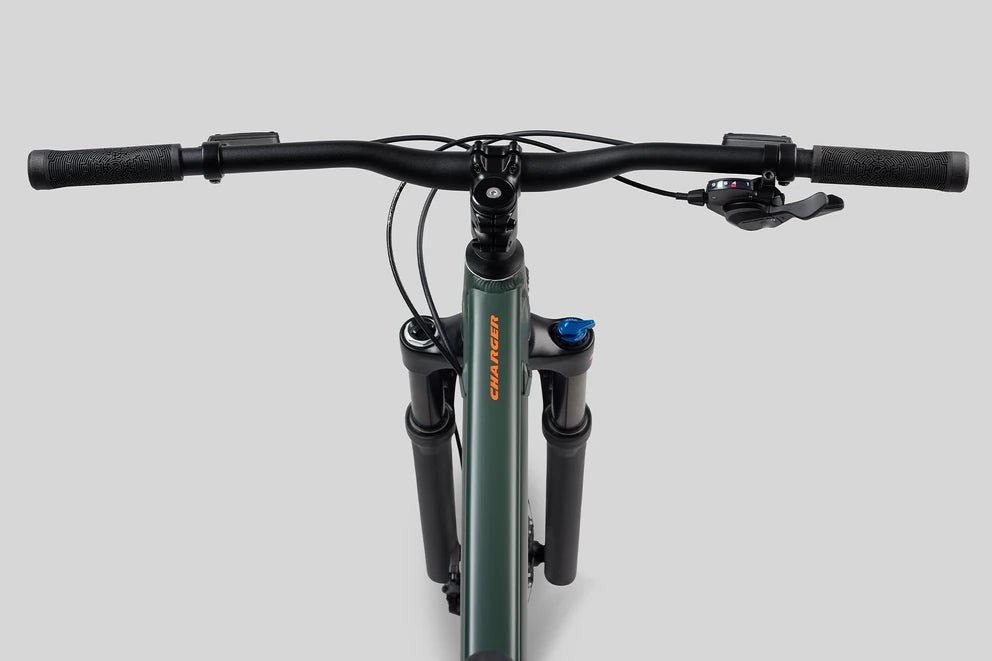 Norco Charger handlebar and front suspension – Top-down view shows black textured handlebars with shifters and brake levers mounted above a dark green bicycle frame and black front suspension fork against a gray backdrop.