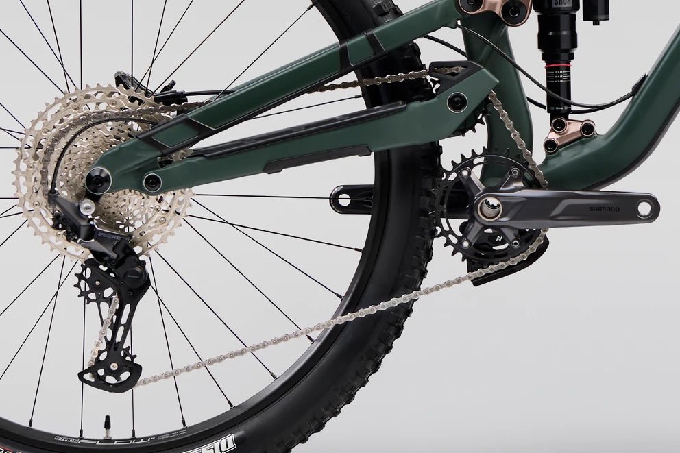 Norco Optic A2 drivetrain and rear suspension – A close-up of a green full-suspension mountain bike highlights the Shimano crankset, rear derailleur, cassette, chain, and rear shock against a plain white background.