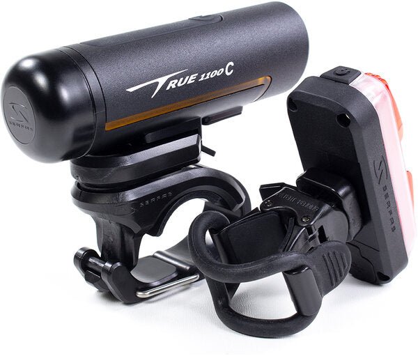 Serfas True Series bike light combo set – A black cylindrical Serfas True 1100C front bicycle headlight sits beside a compact red-lensed tail light, both fitted with black handlebar and seatpost mounting brackets against a white backdrop.